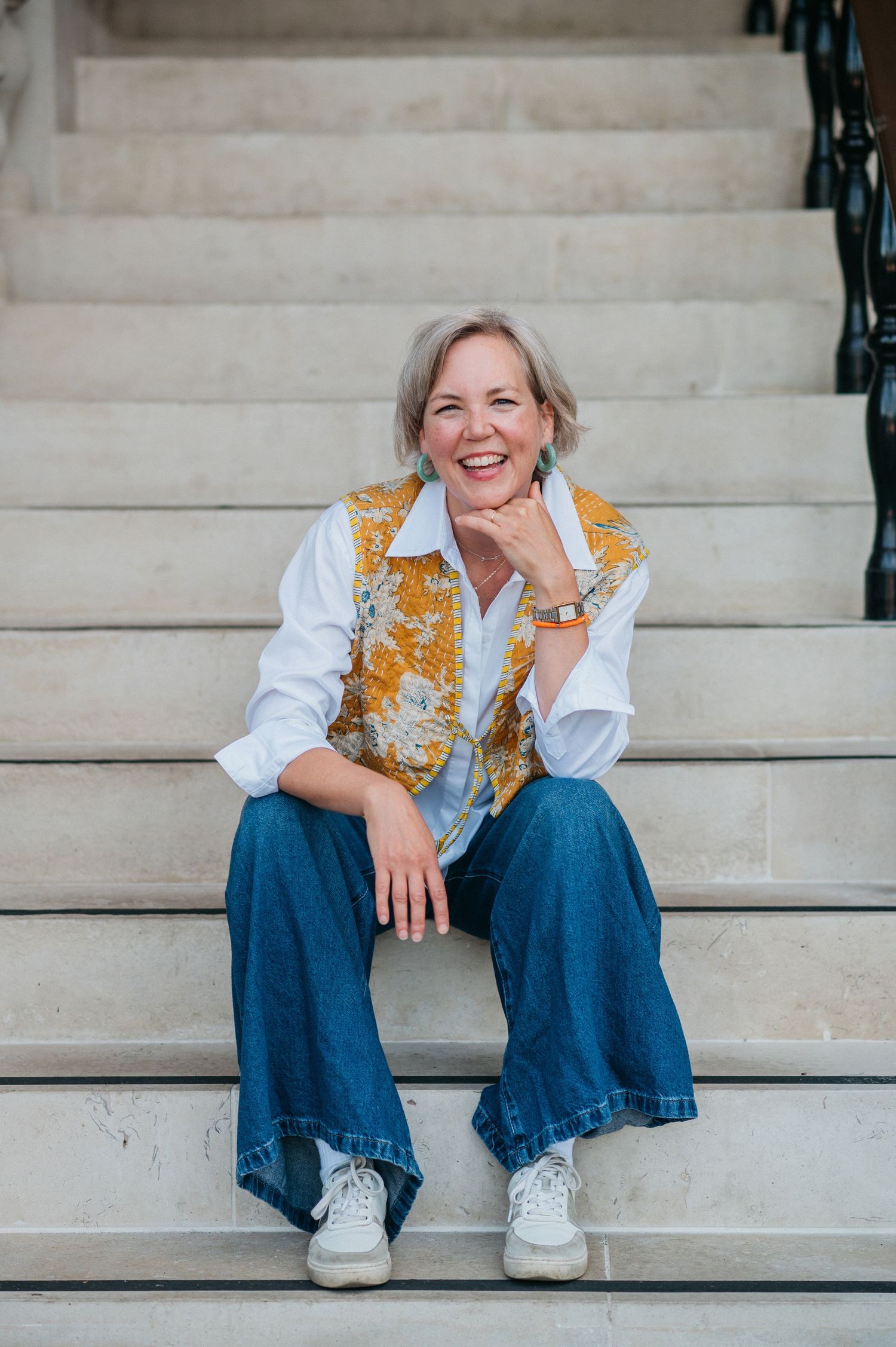 Someone wearing a gold sequined vest and white shirt sits casually on concrete steps while smiling warmly.