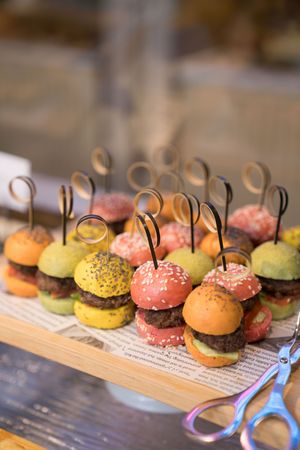 Colorful mini sliders with pink, yellow and orange buns arranged on a wooden board with decorative black picks.色彩繽紛的迷你漢堡，粉紅色、黃色和橙色的麵包，擺放在木板上