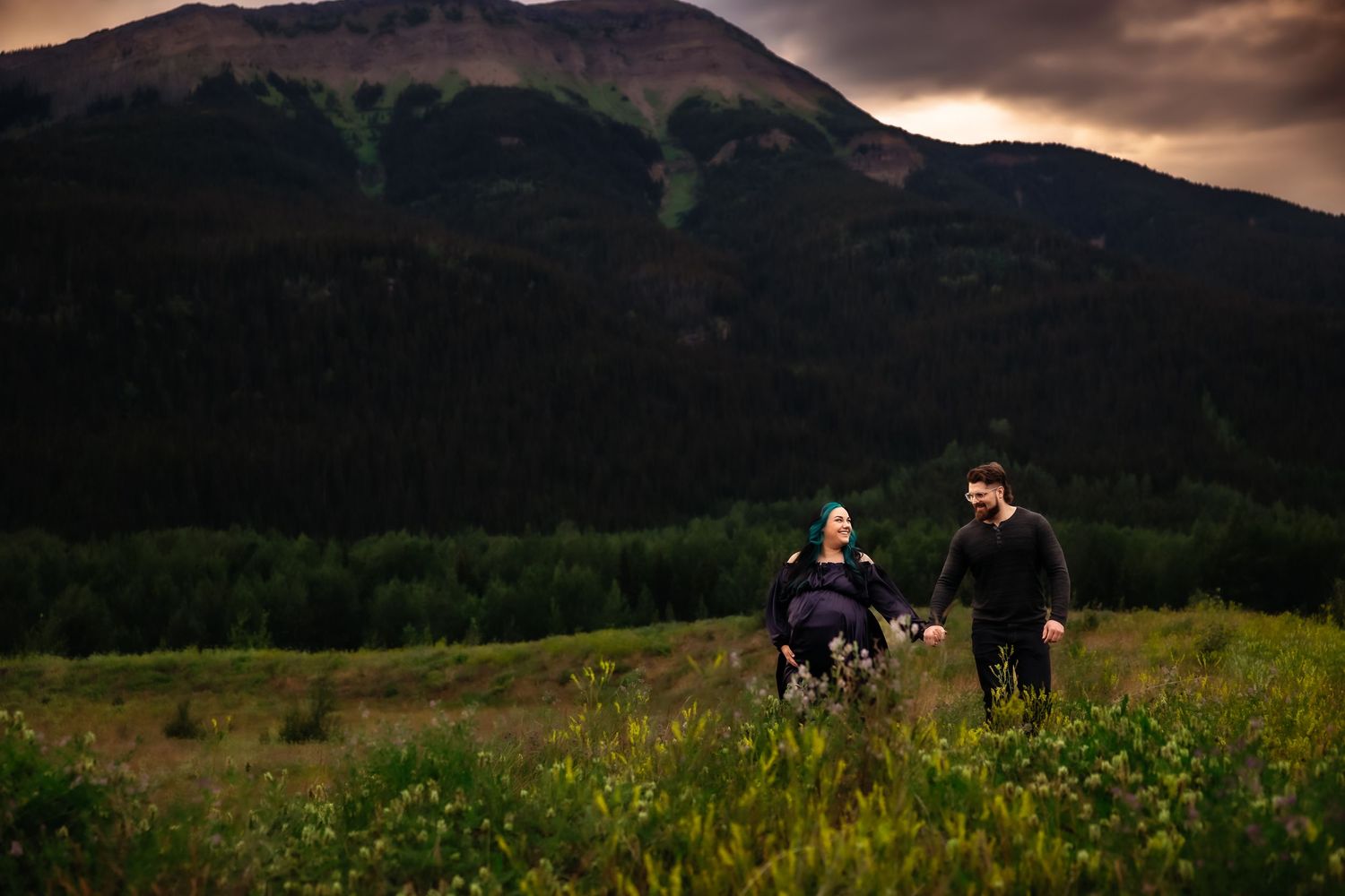 A panoramic mountain landscape at sunset with wildflowers in the foreground and people walking in the distance.