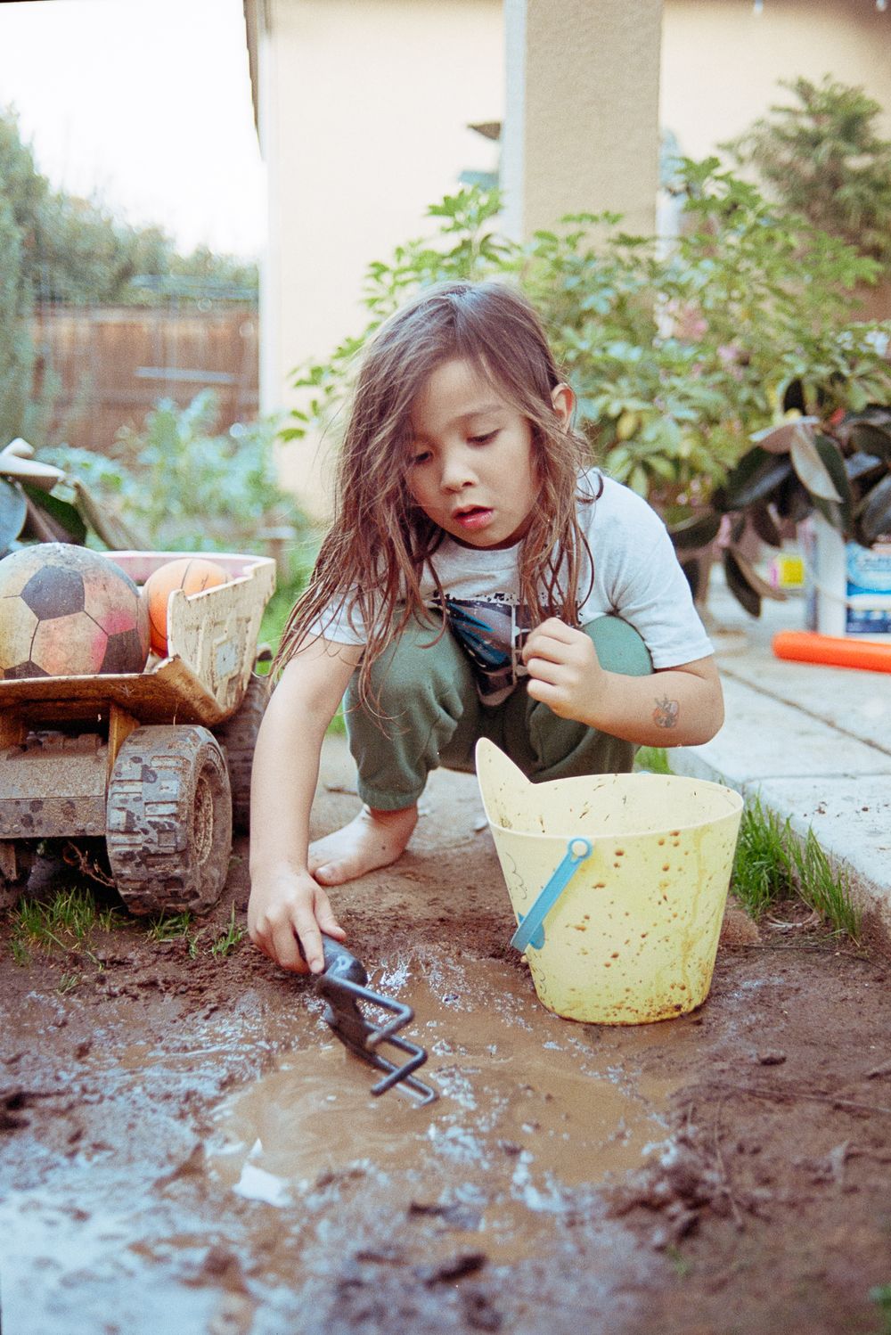 A child in overalls plays in the mud with toys and a yellow bucket in an outdoor setting.