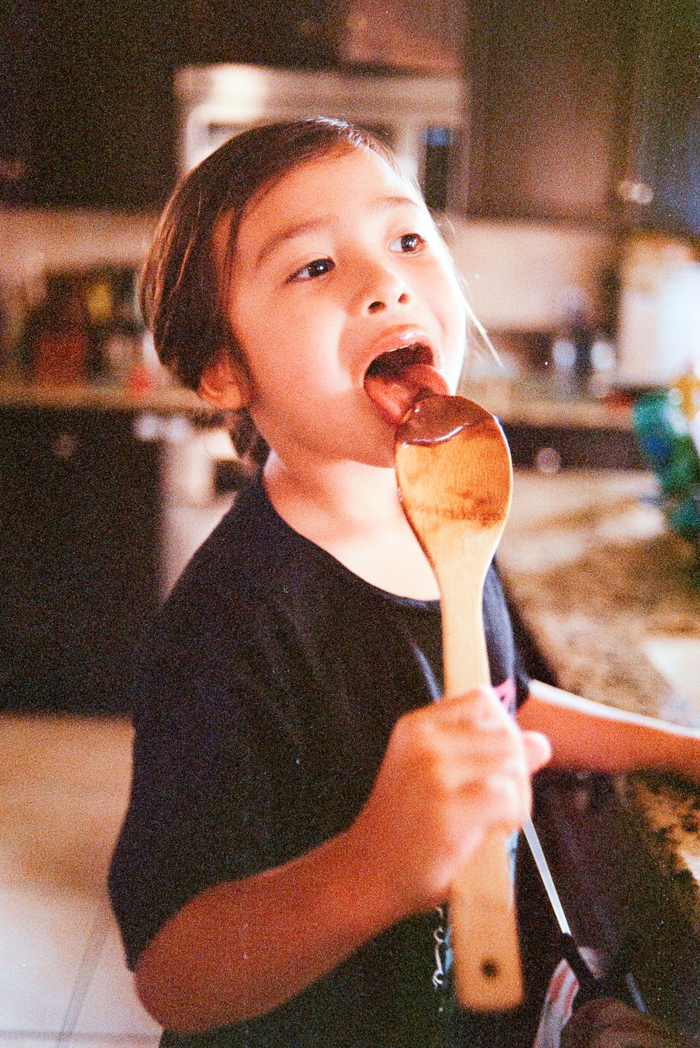 A young child in a black shirt licks cake batter off a wooden spoon in a warm-lit kitchen.