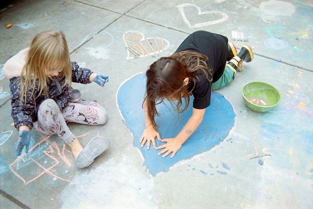Children lay on their stomachs drawing with sidewalk chalk on a driveway covered in heart designs.