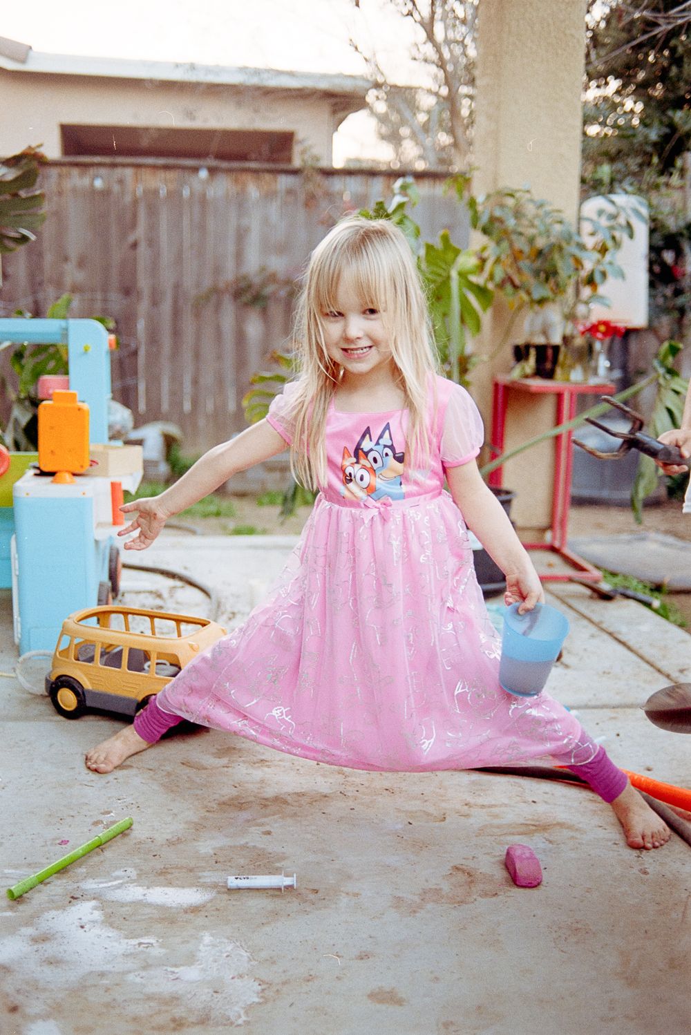 A young child in a sparkly pink dress plays with toys in a backyard patio setting.