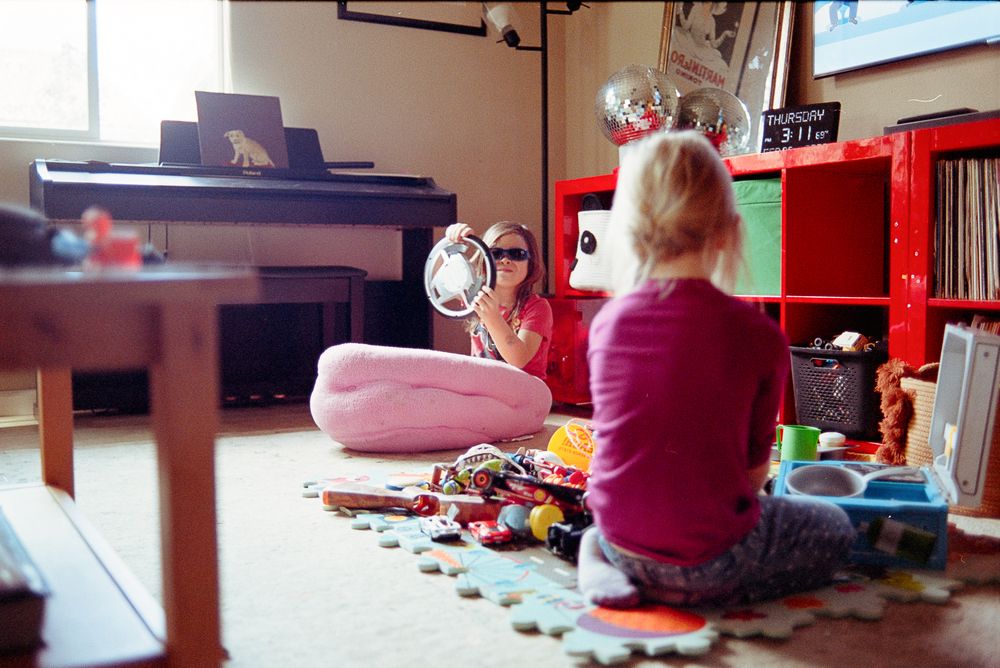 Child in pink sits on colorful play mat surrounded by toys in a living room with piano.