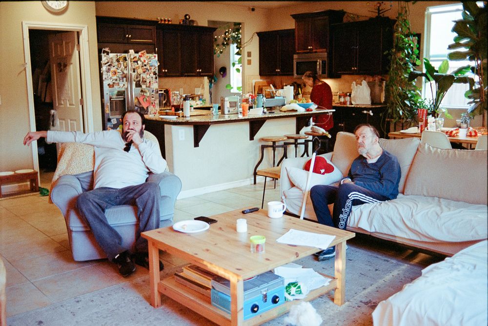 Two people sit on couches in a cozy living room with dark kitchen cabinets visible in the background.