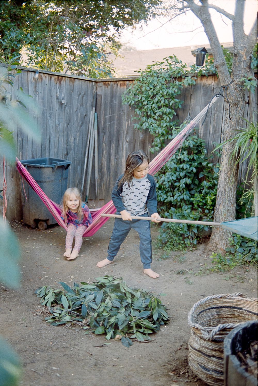 Children play near a wooden fence in a backyard with a pink hammock and lush greenery.