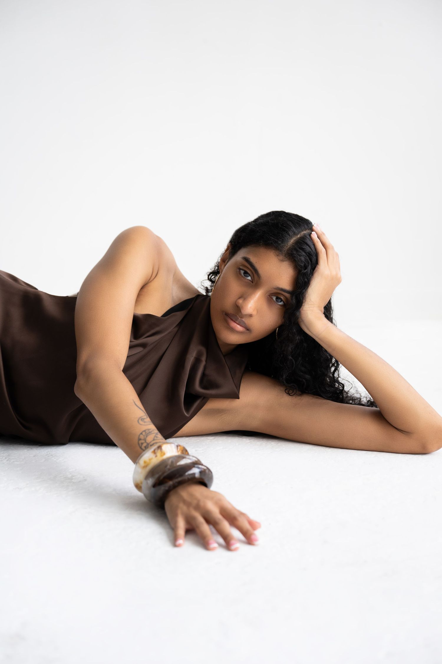 Woman in brown satin dress lying down, wearing a tortoiseshell cuff bracelet and tattoo, posing on white background.