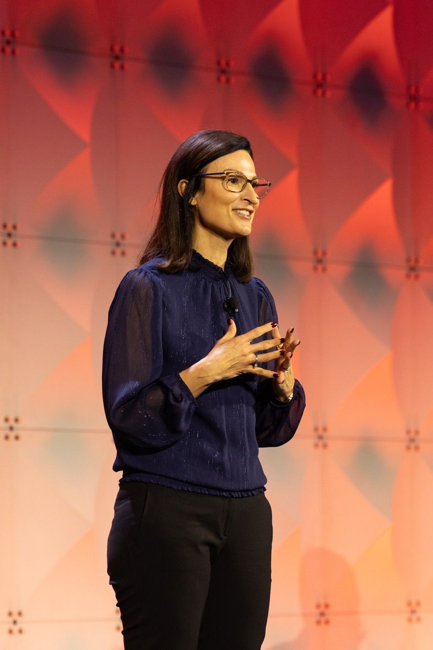 Woman in navy blouse and glasses smiling while speaking on stage with colorful geometric background as the keynote speaker at a Dallas event