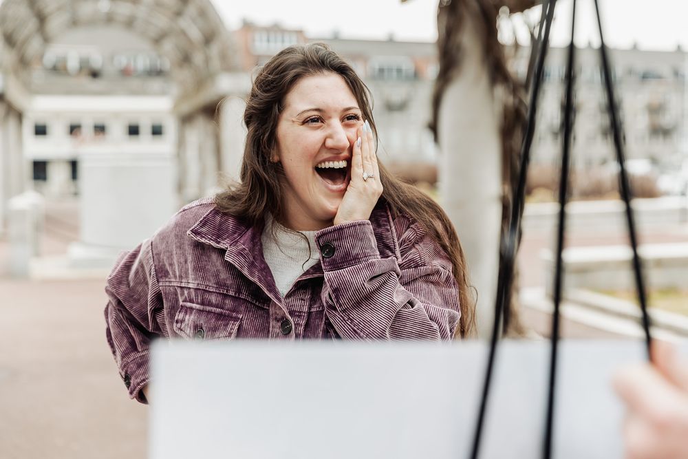 Excited young woman in purple denim jacket laughing outdoors, hand on cheek, holding white bag.