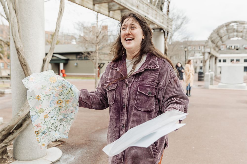 Woman laughing outdoors holding papers near a decorative mosaic sculpture in a public space.