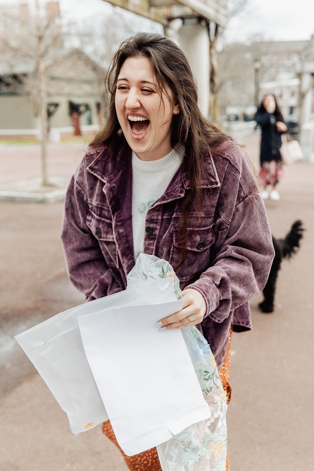 Excited young woman laughing while holding white envelopes outdoors in a park wearing a purple denim jacket.