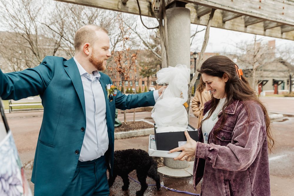 Couple celebrating outdoors with a black dog nearby, man in teal suit gesturing while woman in purple jacket smiles.