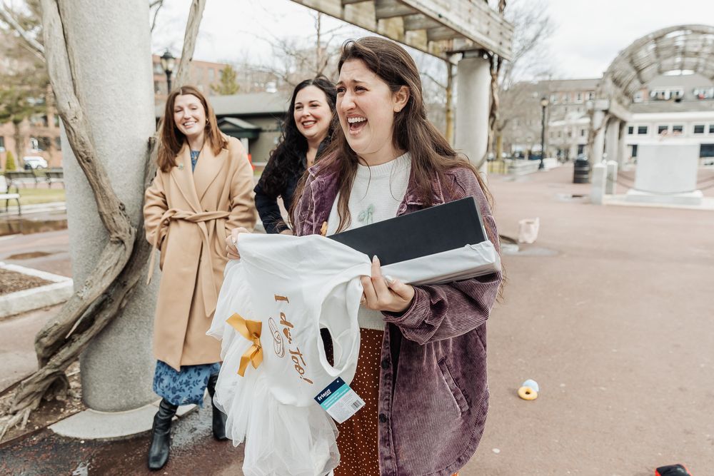 Three young women laughing outdoors as one holds up a white garment, with trees and a plaza in the background.