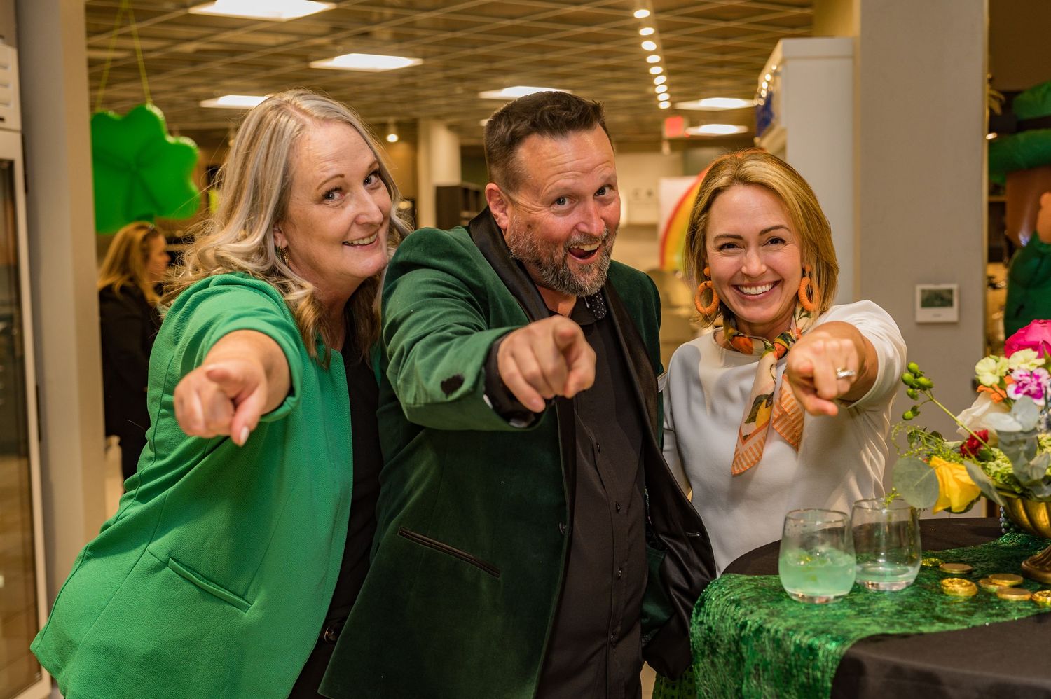 Three smiling people in green outfits pointing at camera at a festive indoor event.