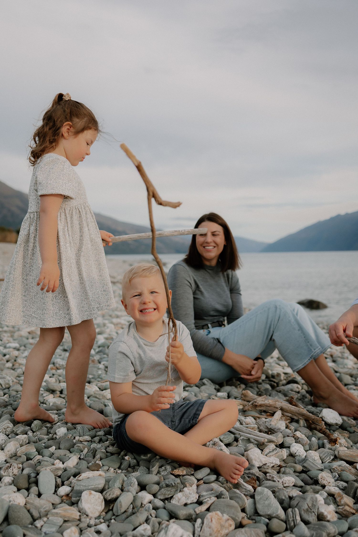 A mother relaxes on a rocky beach while two children play with driftwood, mountains and lake in background during a Queenstown photoshoot.