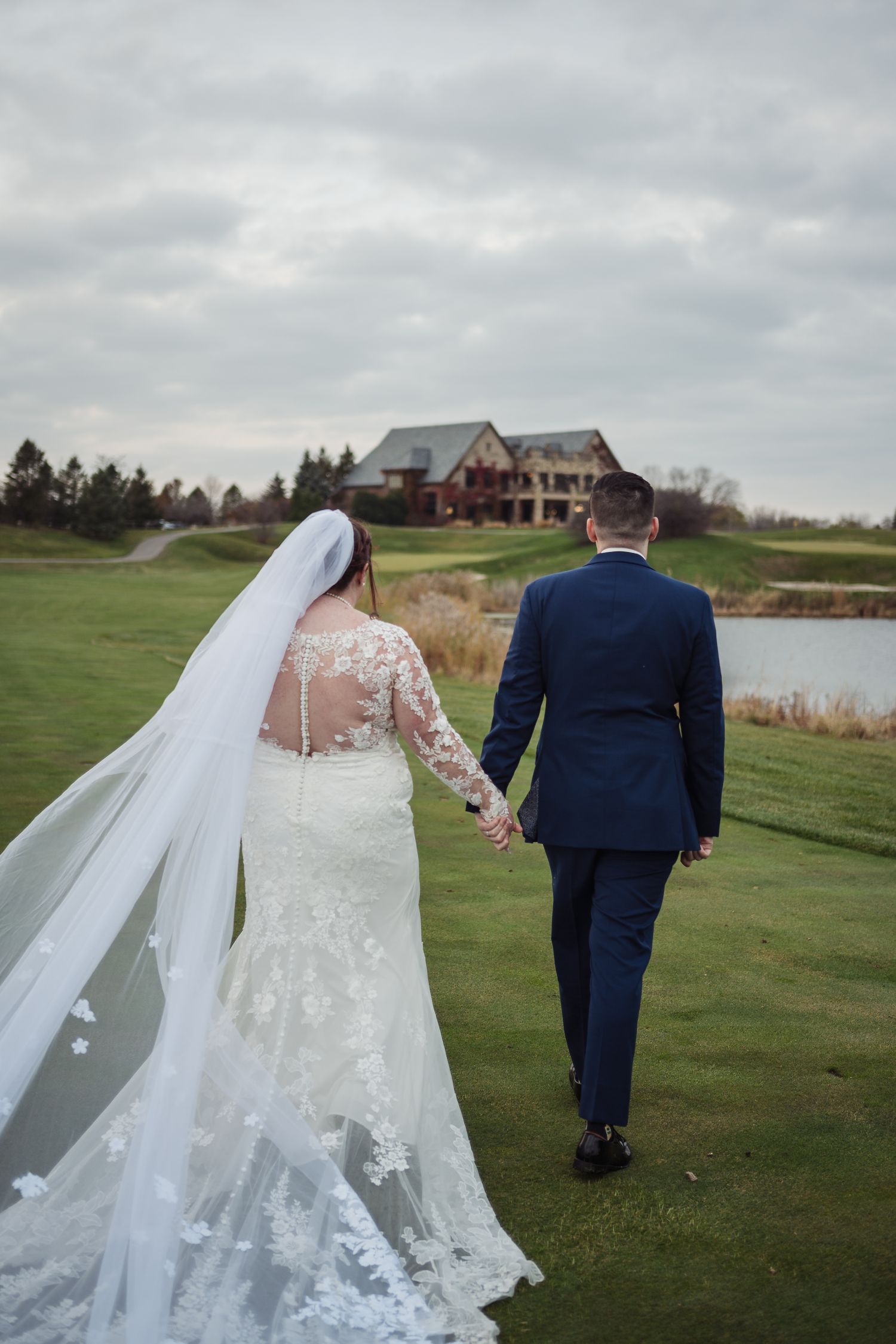 A couple in wedding attire walk hand in hand on a golf course with a rustic lodge in the background.