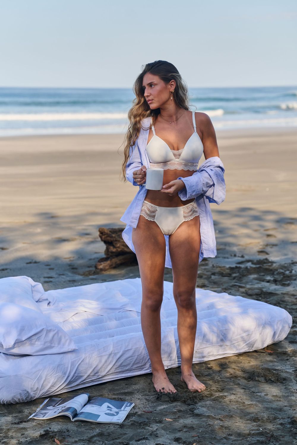 A person in a white bikini and open shirt poses on rocky beach terrain at sunset.