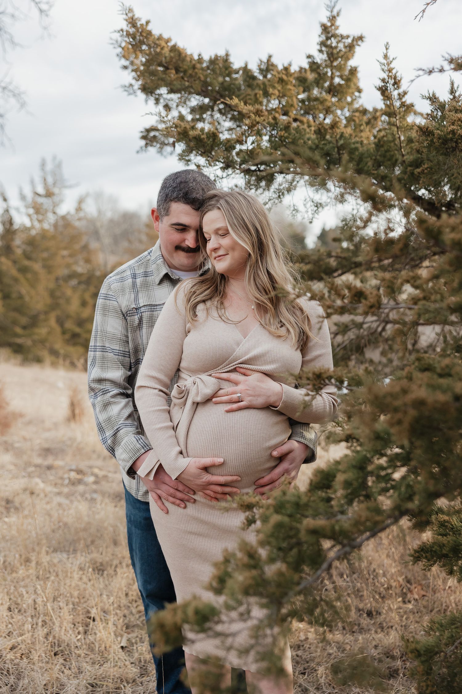 couple stands close together while holding moms baby bump next to pine trees at lake olathe