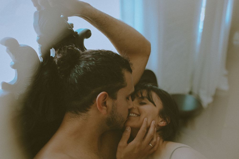 A romantic couple embraces in a dimly lit room with soft lighting and curtains in the background.