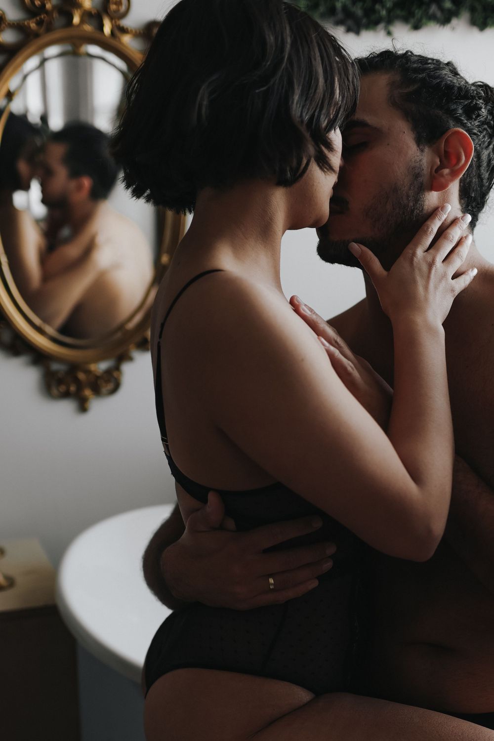 An intimate couple embraces in a dimly lit room with an ornate gold mirror on the wall.