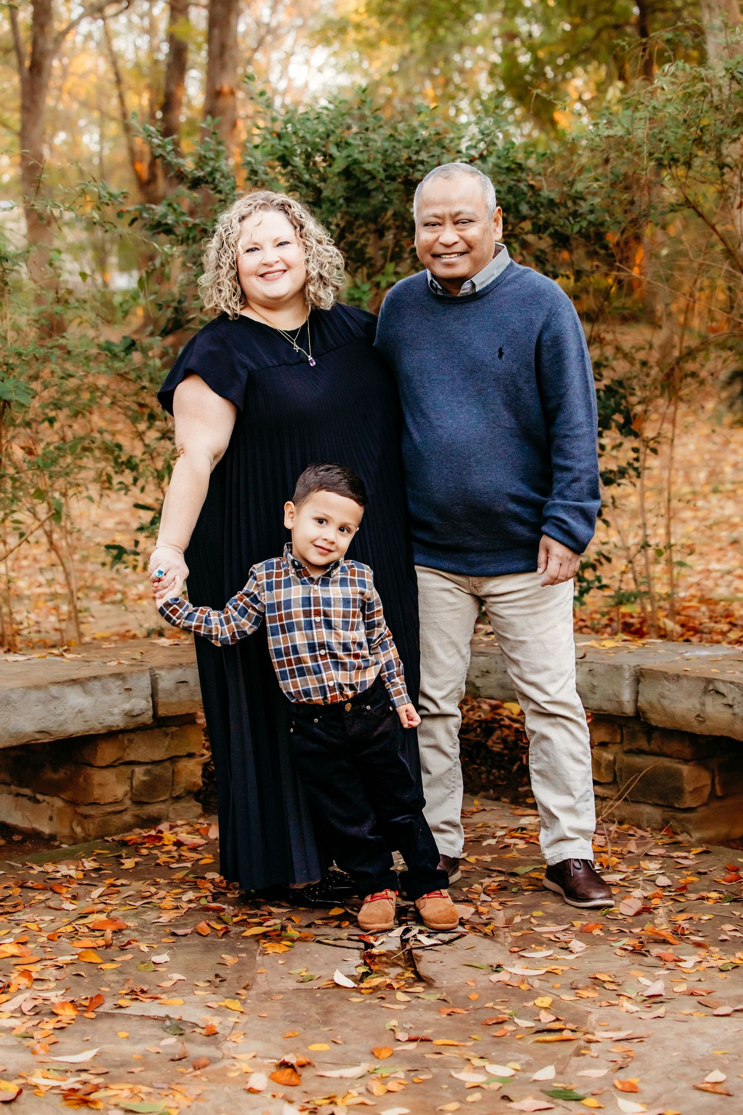 Family of three poses together on stone steps surrounded by autumn foliage in an outdoor portrait setting in Grapevine, TX.