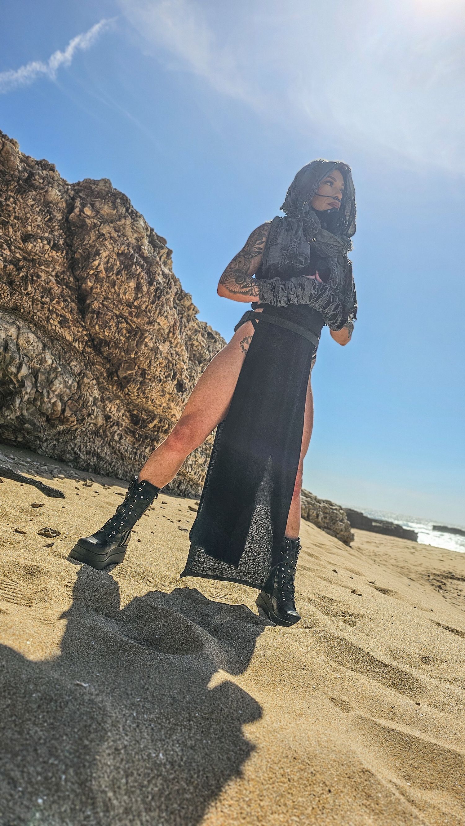 A person wearing a black dress stands on a sandy beach with rocky cliffs and ocean waves in the background.