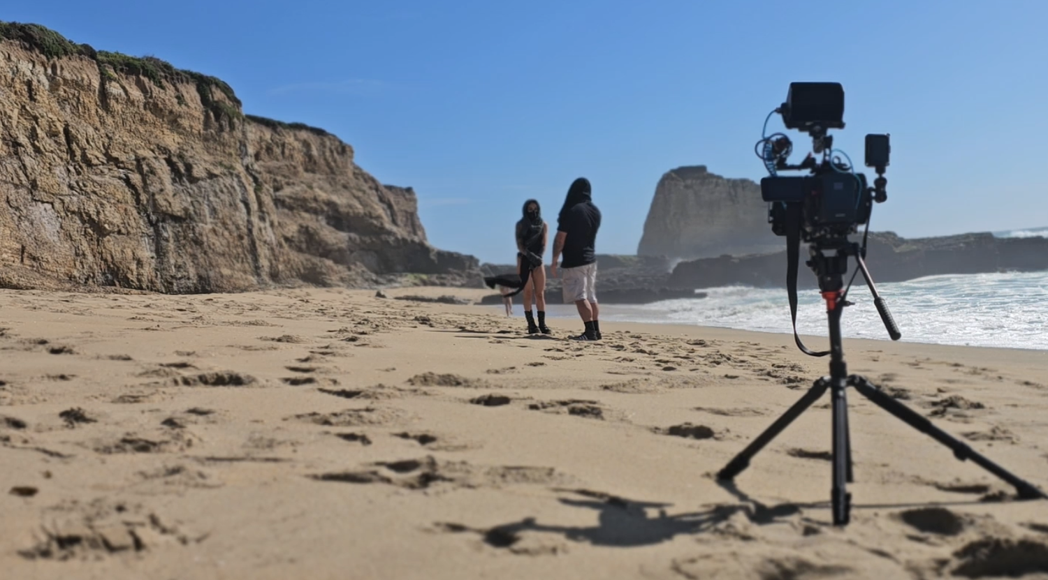 People walking along a scenic coastal beach with dramatic cliffs and rocky outcrops in the background.