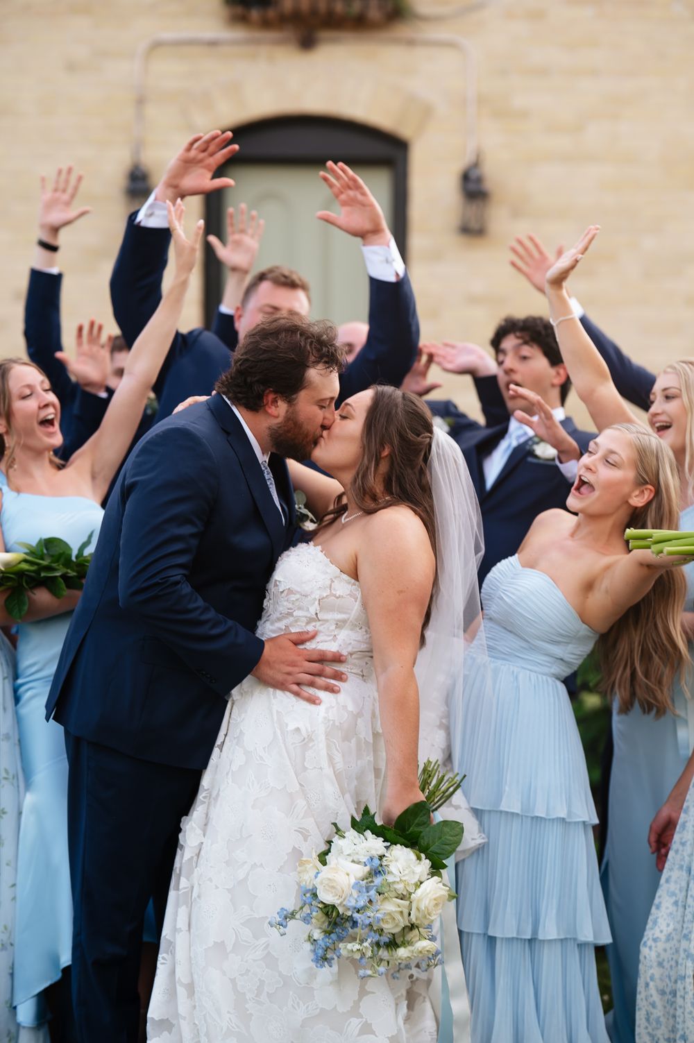 Wedding party in light blue bridesmaid dresses and dark suits celebrating joyfully with raised arms outdoors.