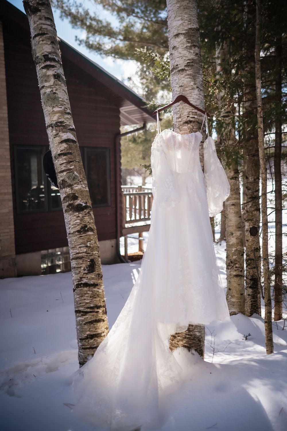 Snow drifts form tall peaks against birch tree trunks near a wooden cabin in a winter forest setting.