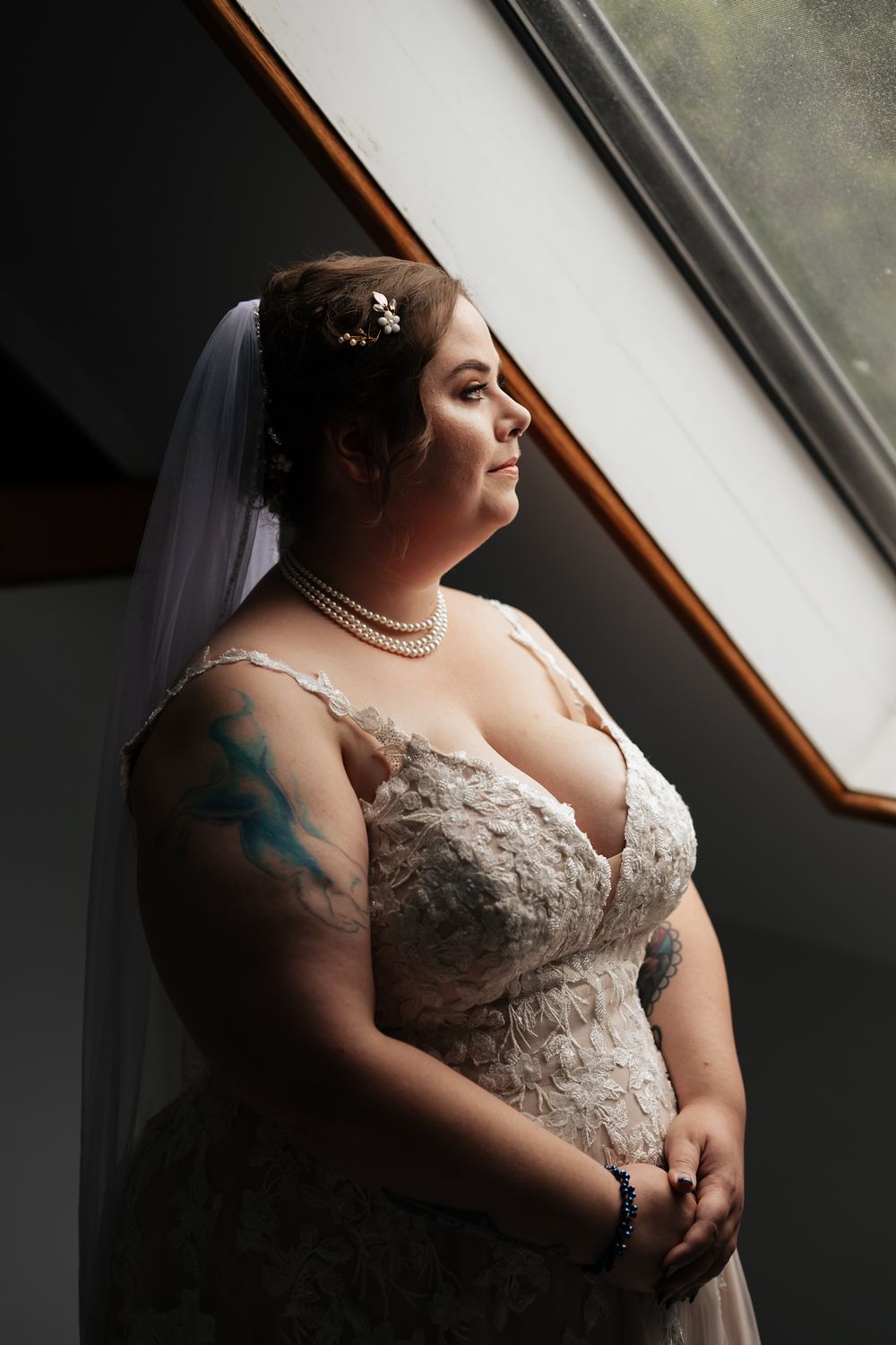 A bride in a lace wedding dress with tattoos stands by a window in soft natural light.
