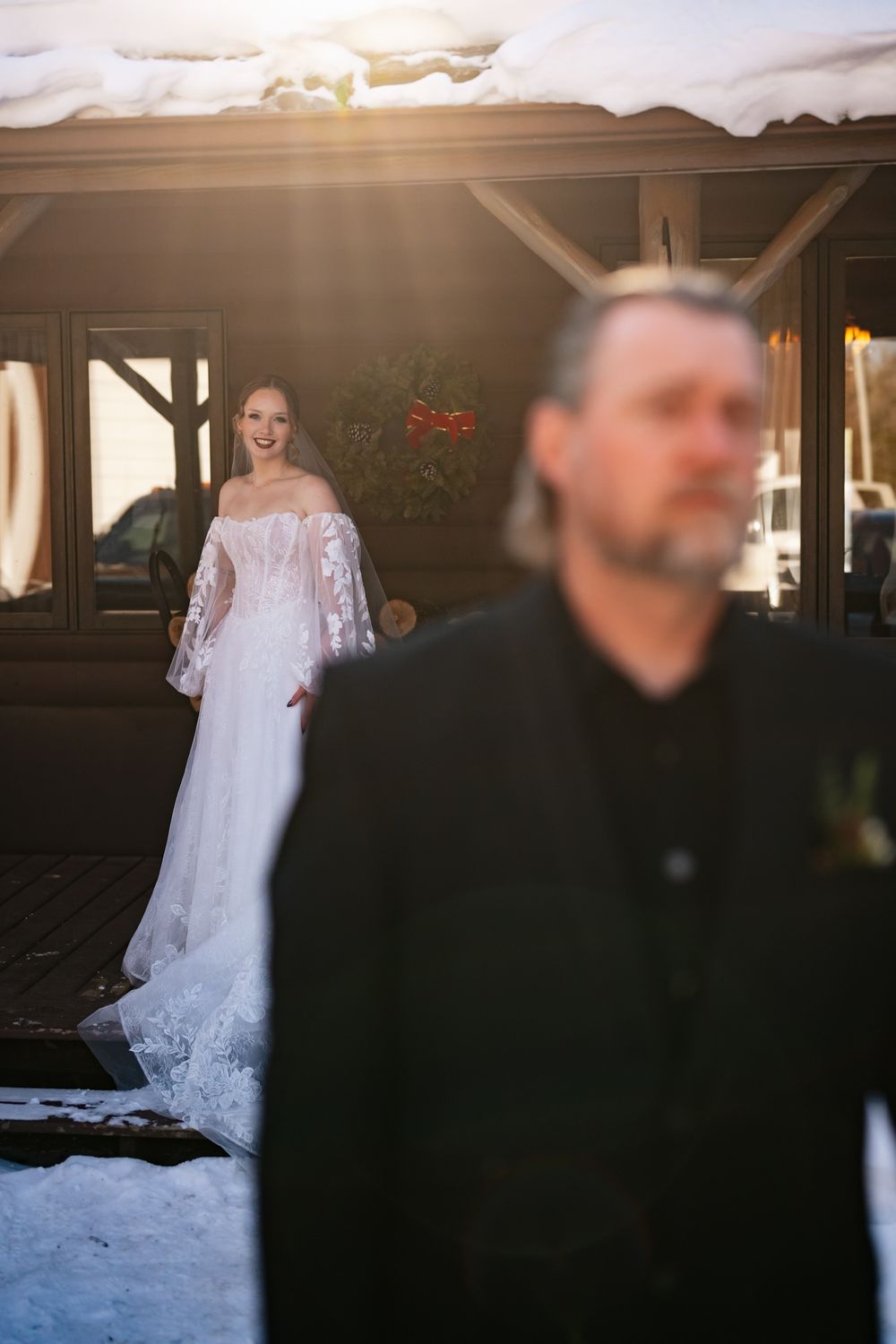 A bride in a white lace wedding dress with long sleeves stands in the background while a silhouetted figure is in the foreground.