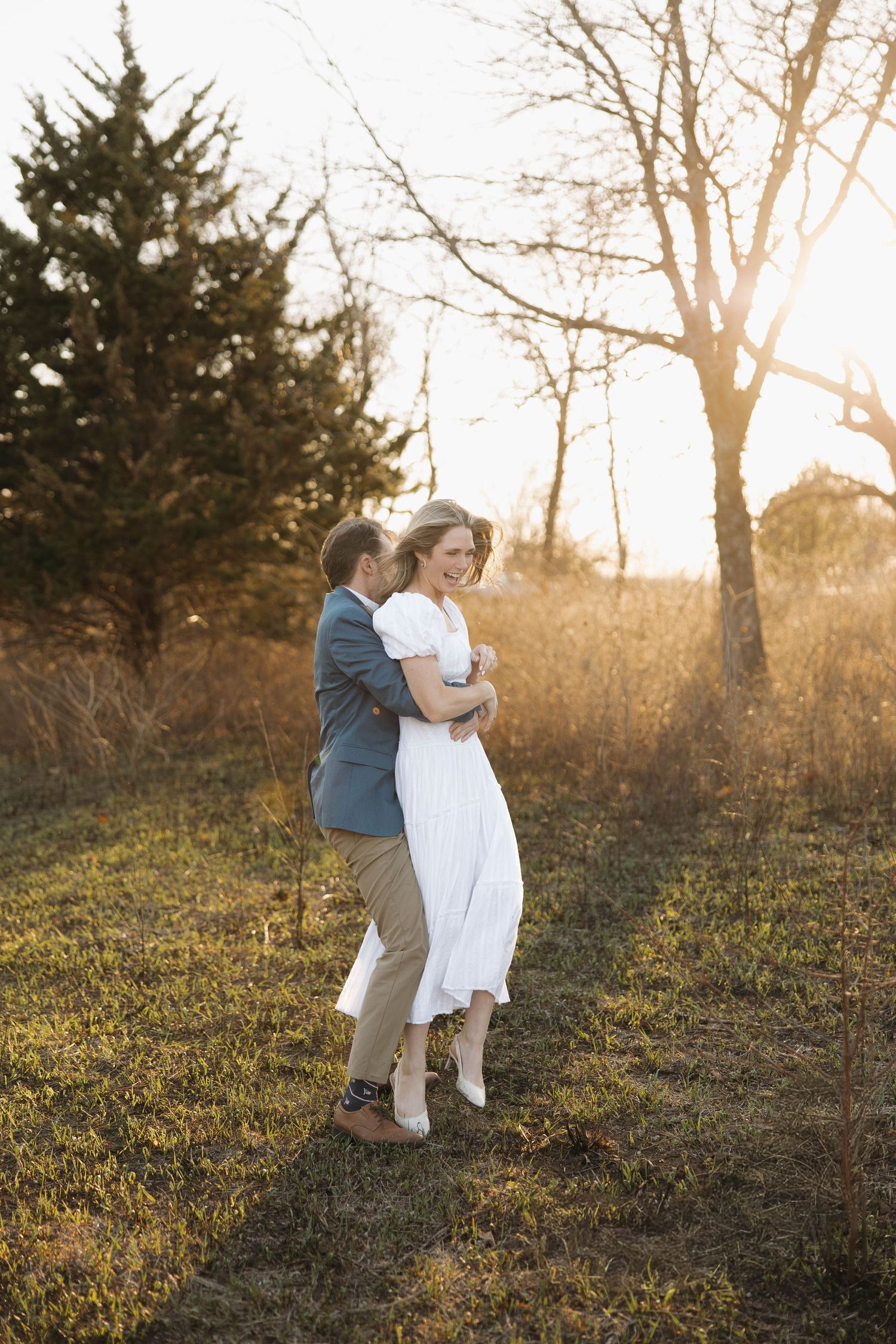 A couple embraces in a golden sunset field with evergreen trees and bare branches silhouetted in the background.