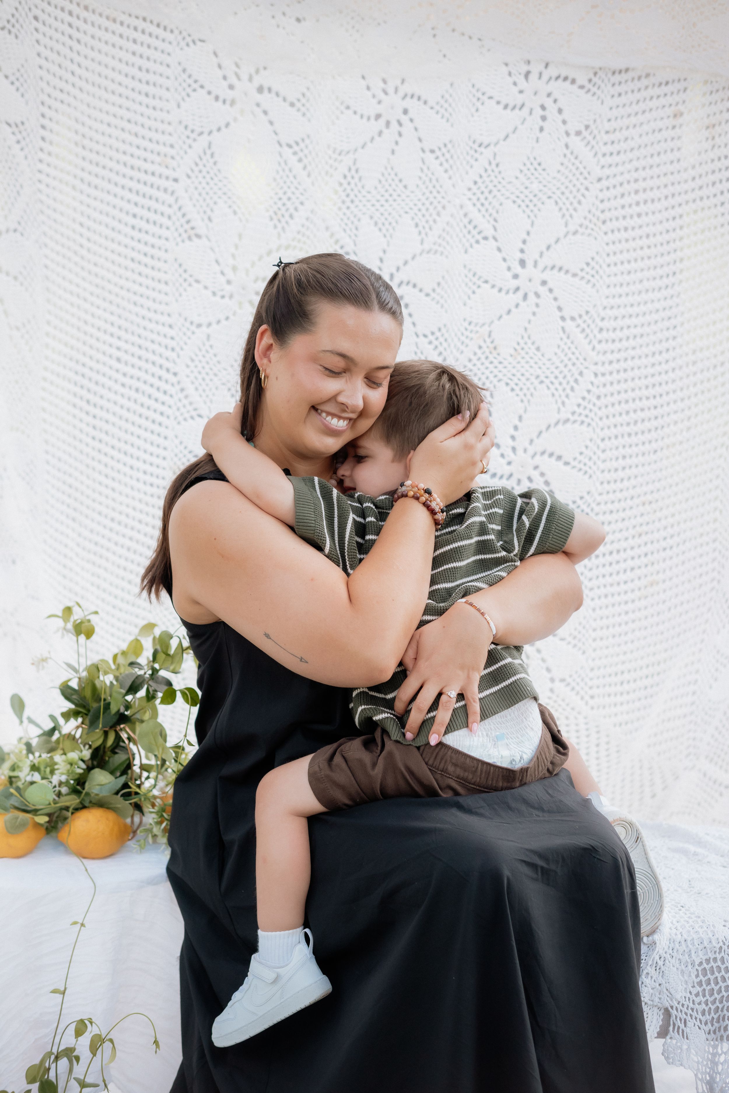 Mother and child share tender moments on white lace backdrop with citrus and greenery decor.