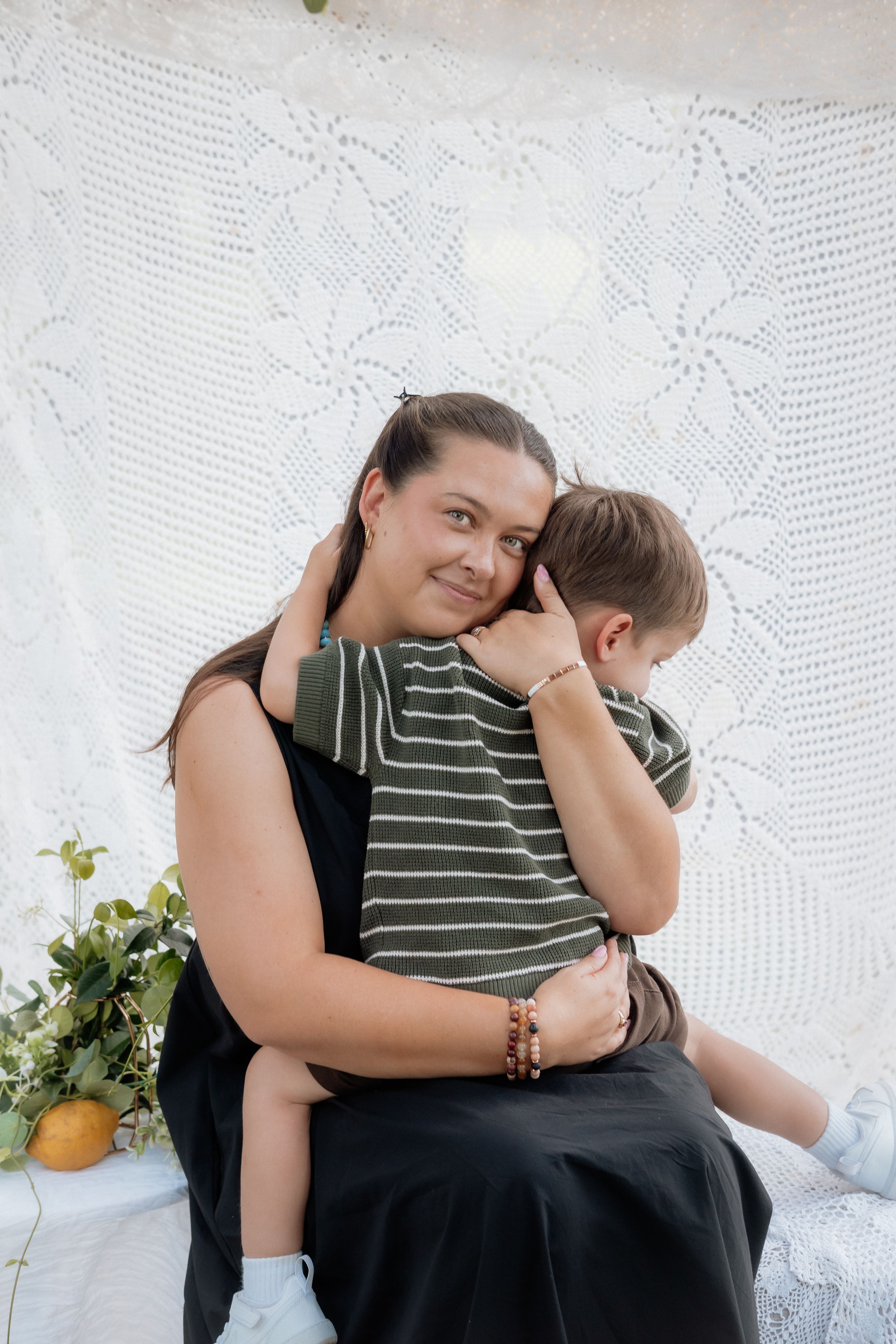 A tender moment shared between parent and child sitting together against a white lace backdrop with citrus fruits nearby.