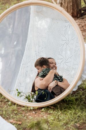 A mirror reflection captures an intimate embrace between parent and child against a lace background outdoors.