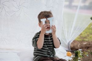 Young boy in striped shirt holding up playing cards while sitting by white curtains outdoors.