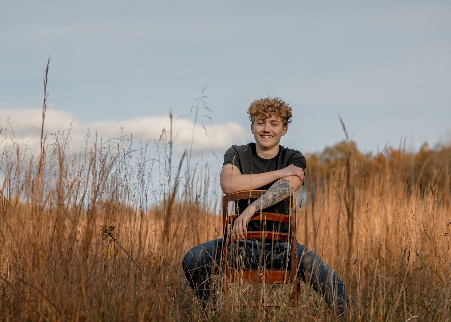 A person sits in tall golden grass at sunset wearing dark clothing and boots.