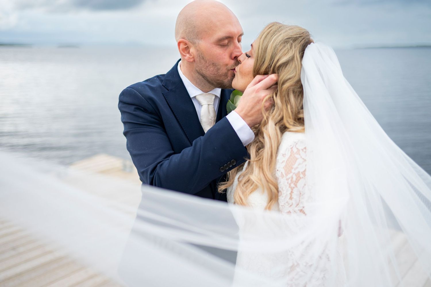 Bride and groom sharing a romantic kiss on a waterfront dock, bridal veil flowing in the wind.