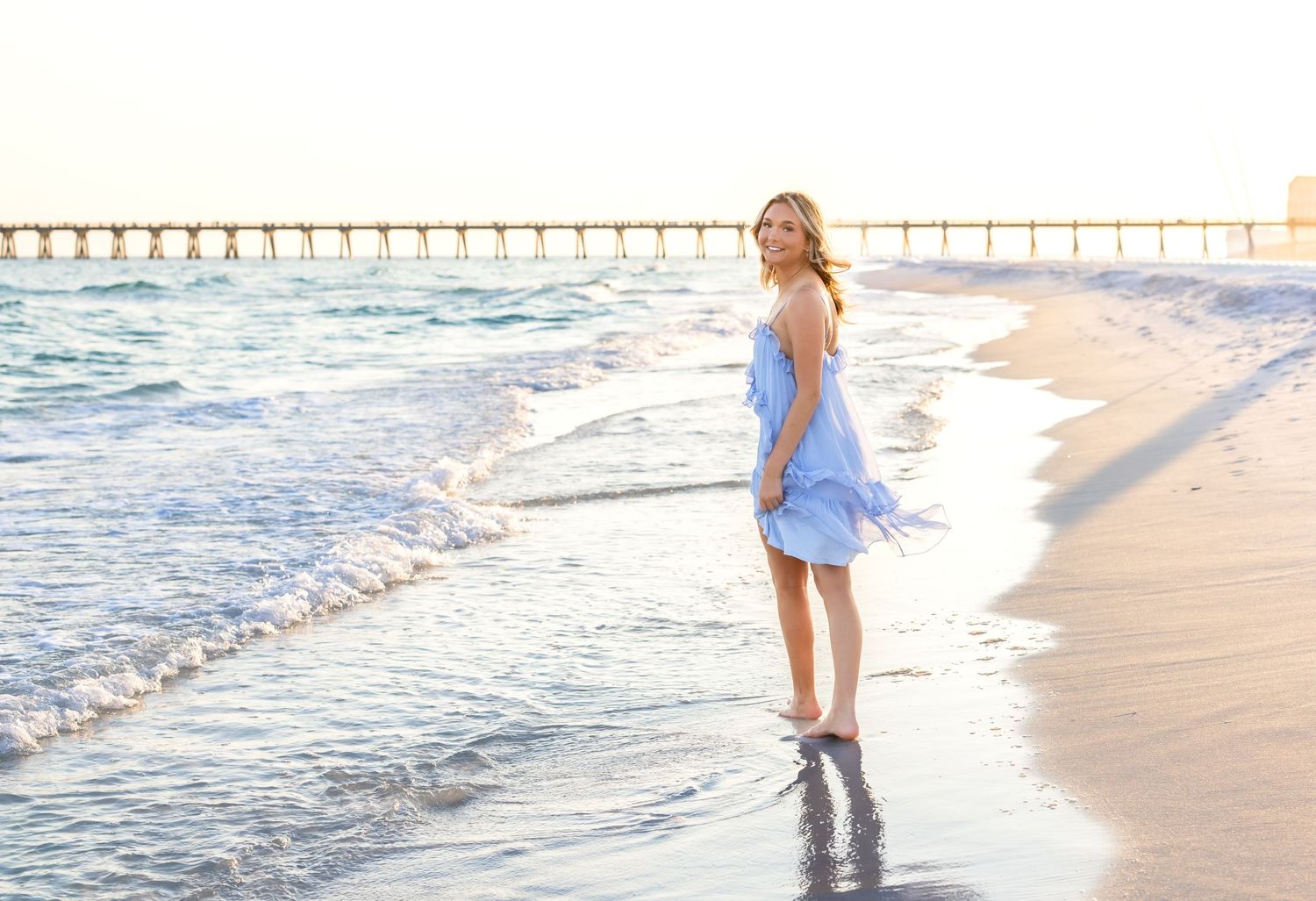 Young woman in blue dress walking barefoot on a sunny beach with waves and a pier in the background.