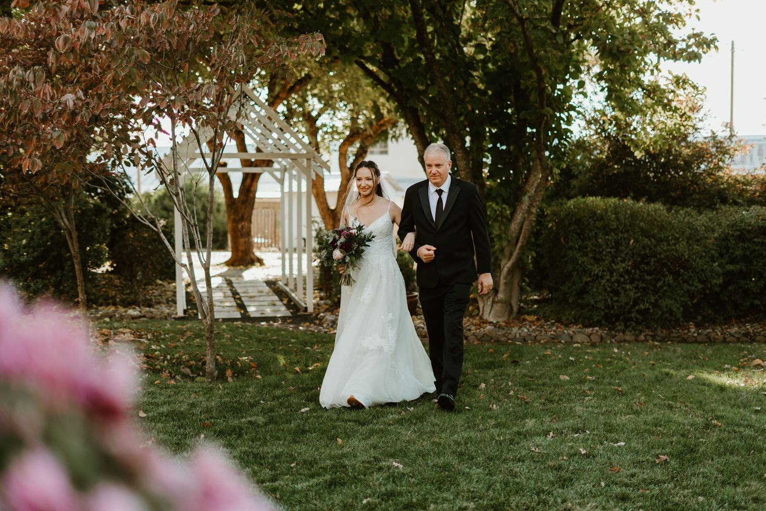 Bride in white gown walks with suited man through lush garden setting on a sunny wedding day.