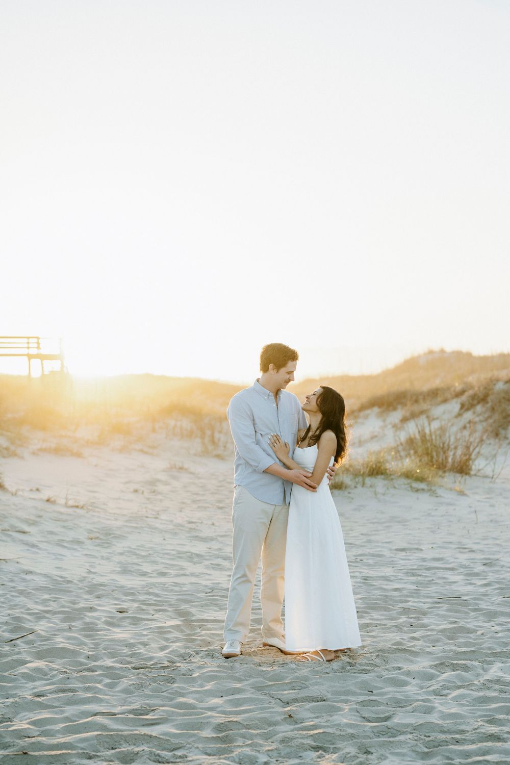 Couple embracing on sandy beach at golden sunset, woman in white dress and man in light blue shirt.