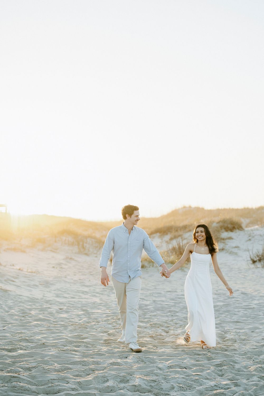 Couple holding hands walking on sandy beach at golden sunset, man in blue shirt and woman in white dress smiling.