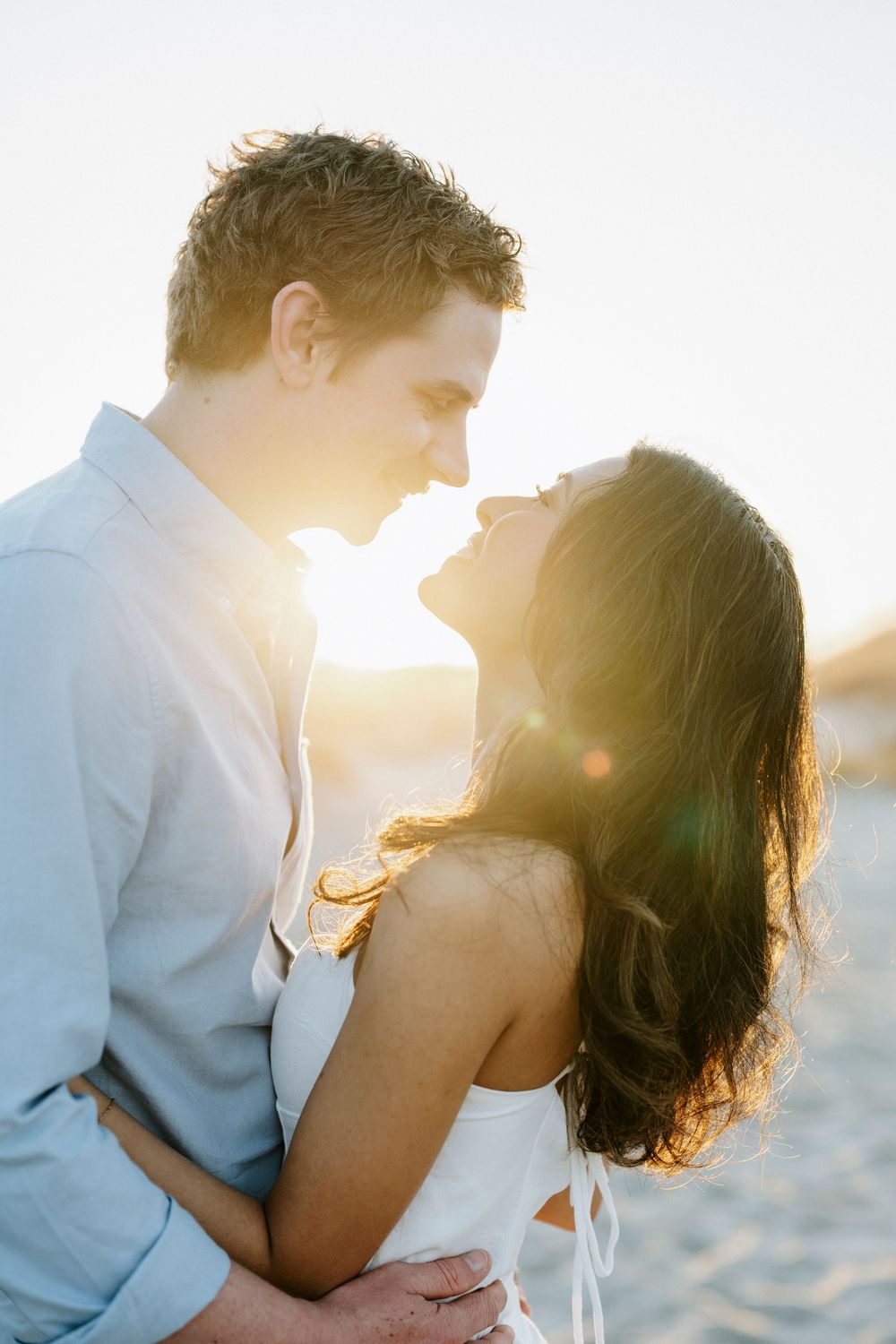 Romantic couple almost kissing at golden sunset on beach, man in blue shirt holding woman in white dress, sun flare.