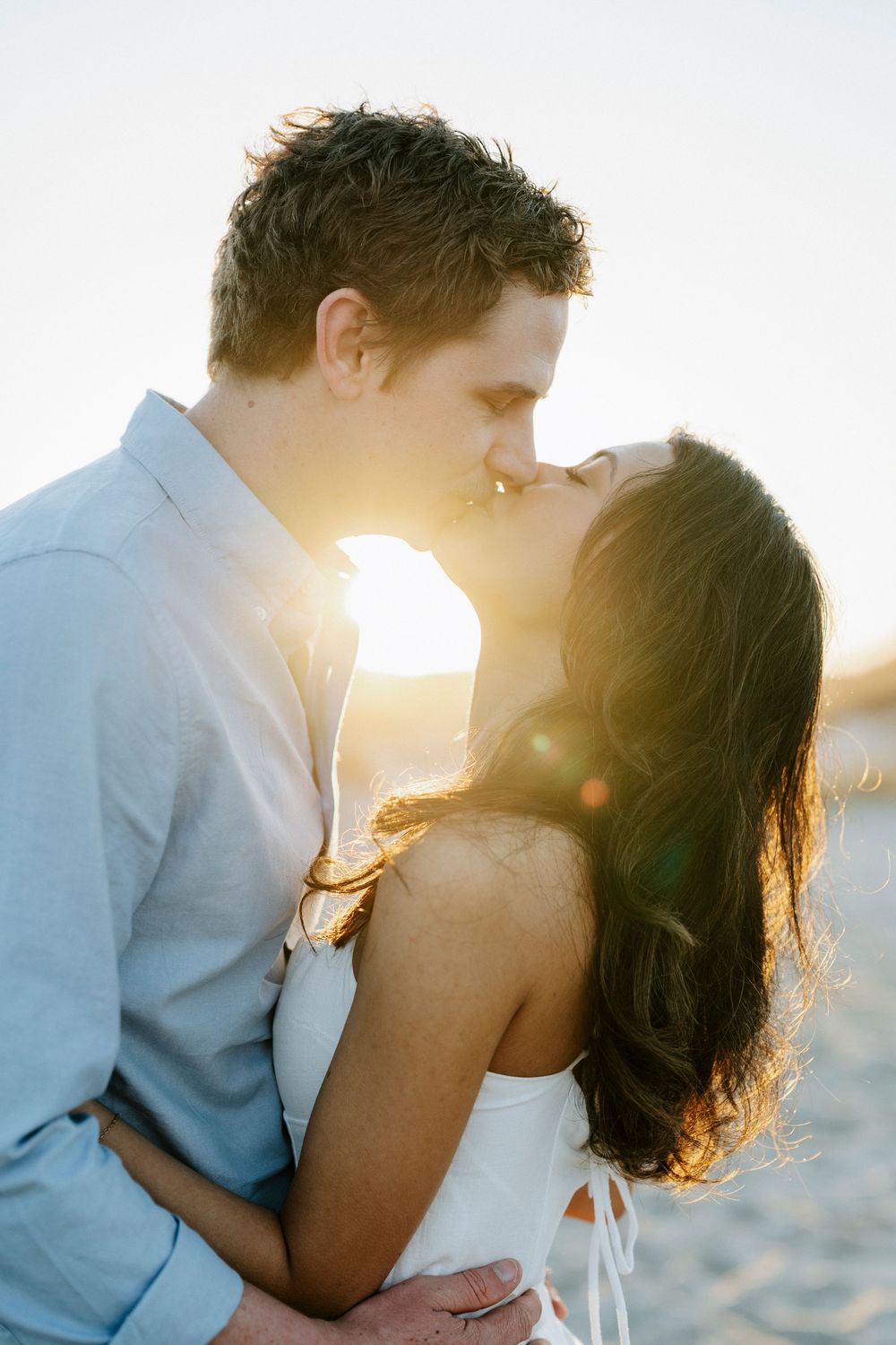 Couple sharing a kiss on the beach at golden hour with warm sunlight glowing behind them.