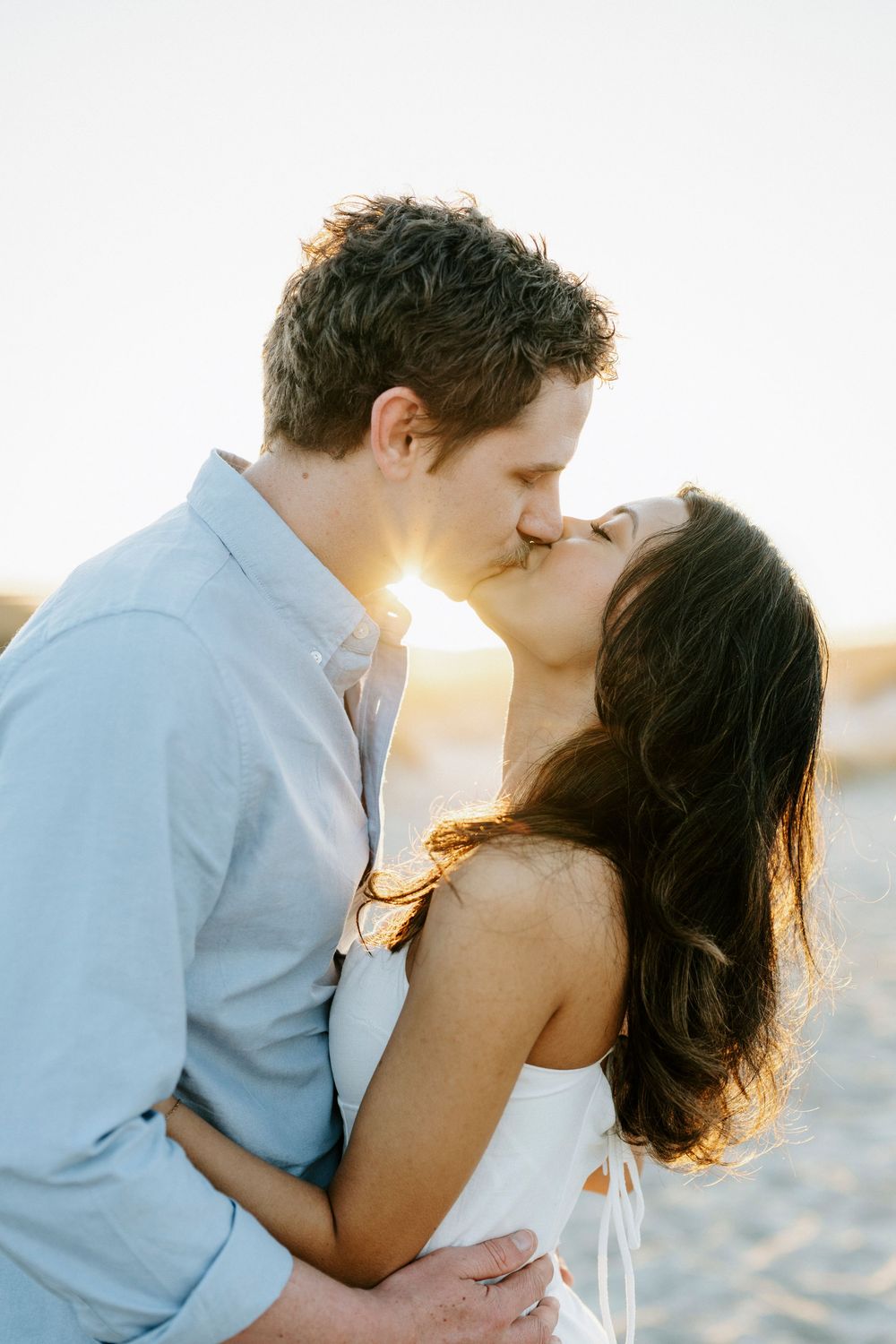 A couple shares a romantic kiss on the beach at sunset, with golden backlight illuminating their embrace.