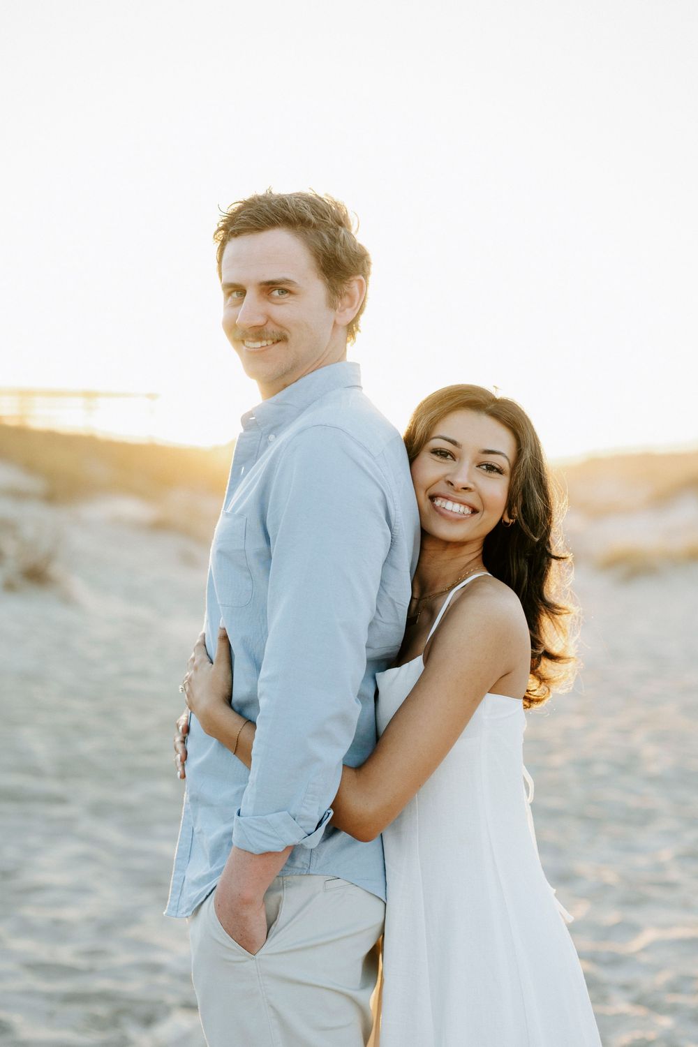 A couple embraces on a sandy beach at golden hour, the woman in a white dress hugging a man in a light blue shirt.
