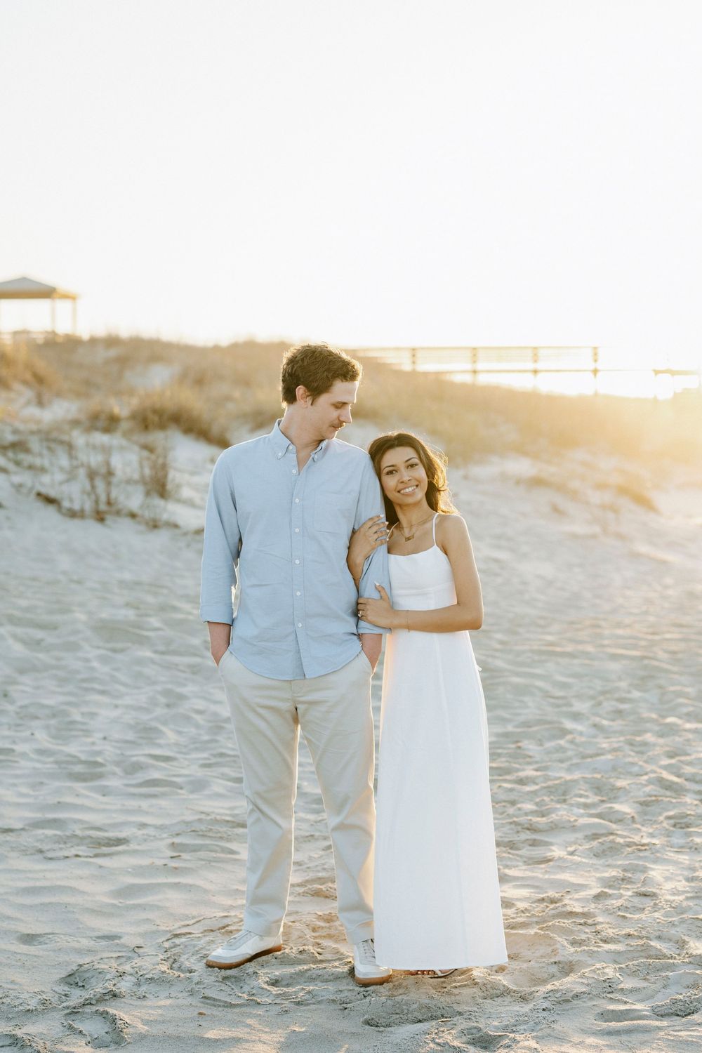 Couple embracing on sandy beach at golden sunset, woman in white dress and man in light blue shirt.