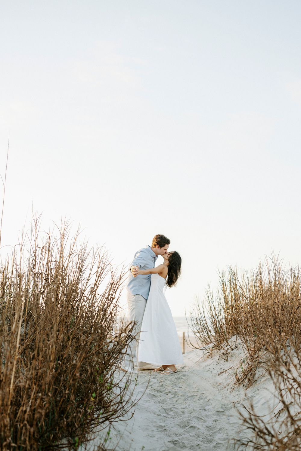 Couple sharing a romantic kiss on sandy beach dunes surrounded by tall coastal grasses at sunset.