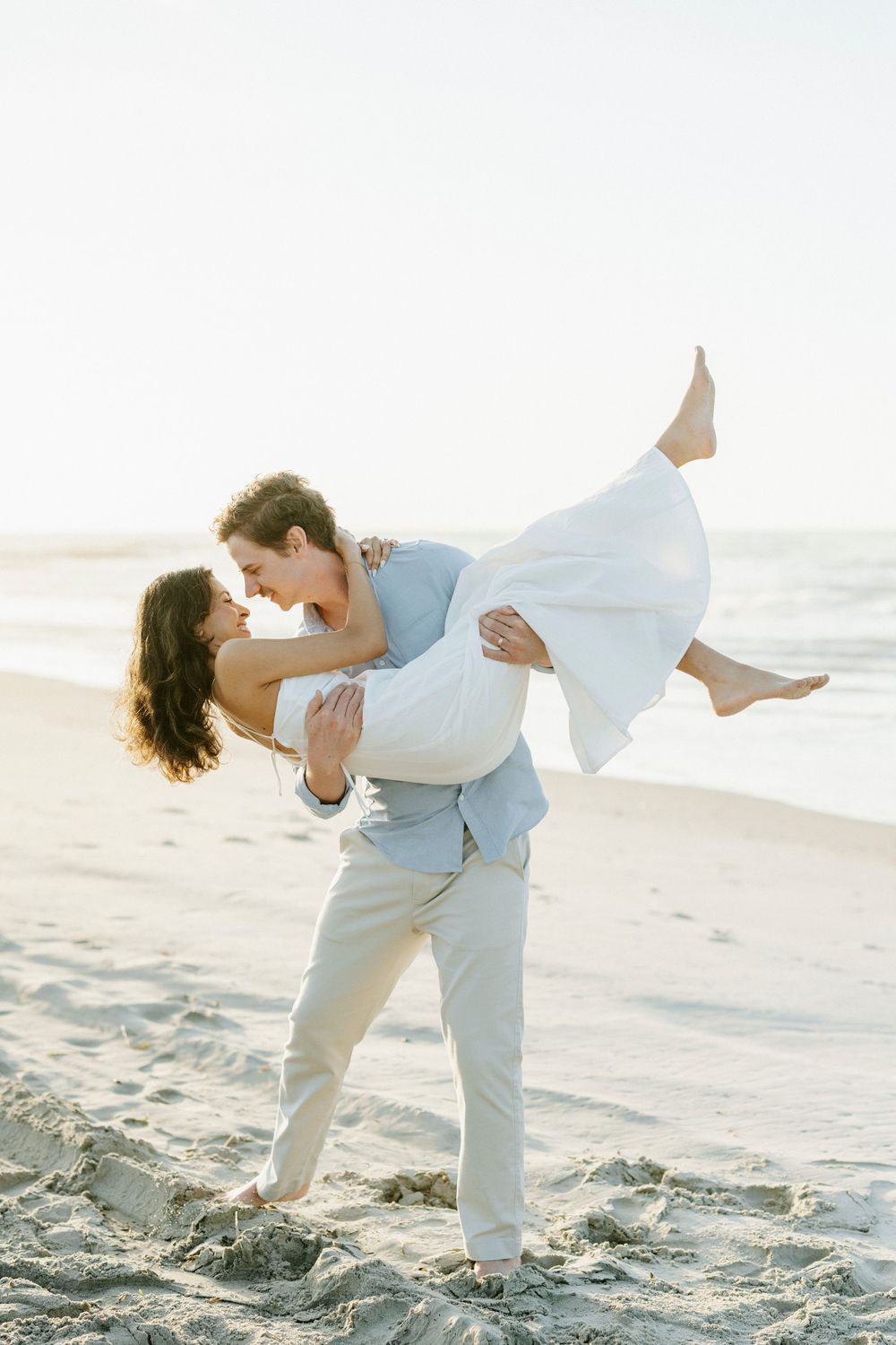 Groom lifting bride on beach, sharing romantic kiss at sunset in light blue suit and white wedding dress.