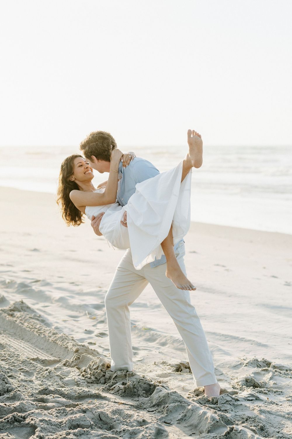 Man lifting woman in white dress on beach at sunset, both laughing joyfully near ocean waves.